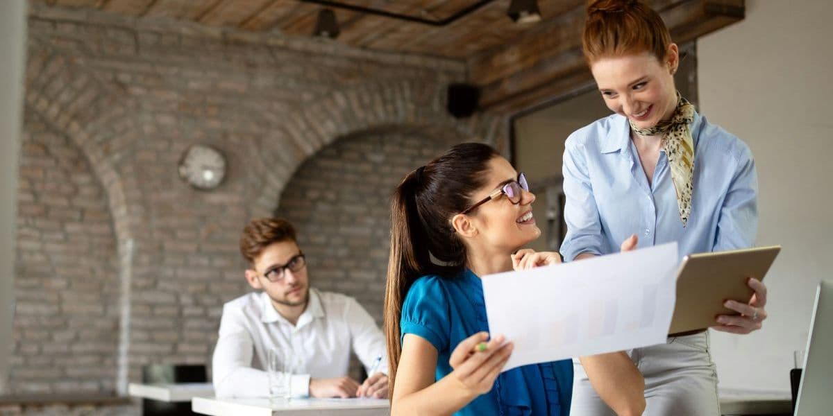Student showing a paper to a teacher in a classroom while another student sits behind.