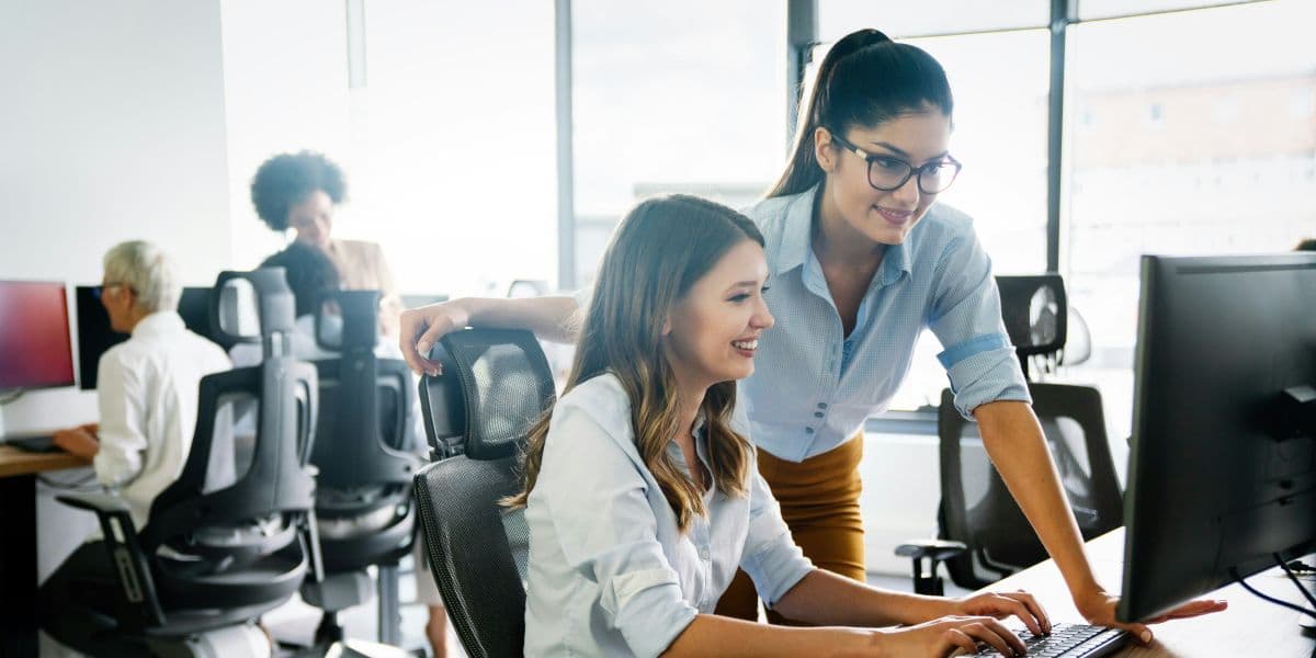 wo women collaborating at a computer in a workplace, with one showing something on her screen to the other, while colleagues work nearby.